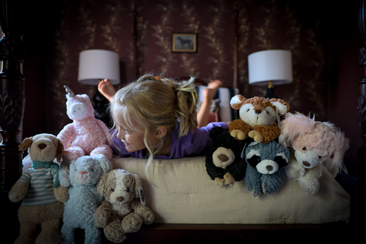 A young girl with blonde hair in a ponytail lies on a bed surrounded by a collection of plush stuffed animals, including a pink unicorn, a bear, a puppy, and a raccoon, in a dimly lit bedroom.