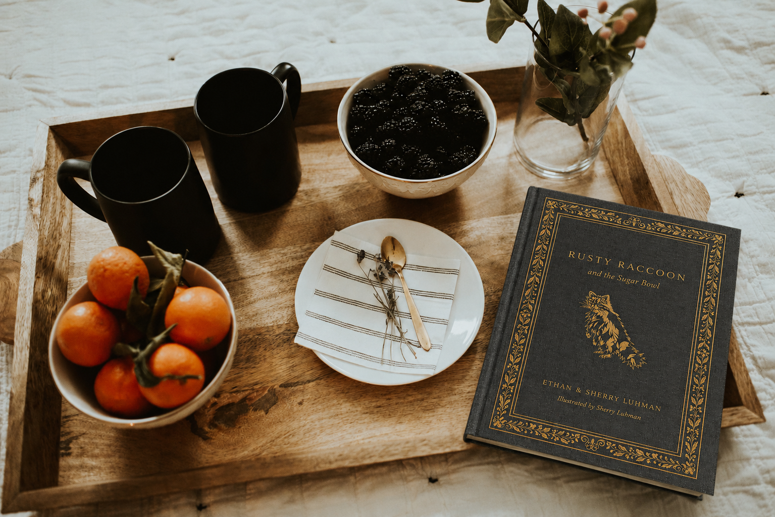 Wooden tray with mugs, oranges, a bowl of dark food, a vase, and a book on a light wooden surface.