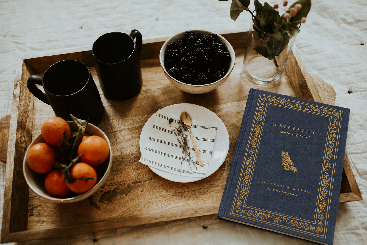 Wooden tray with a bowl of oranges, black mugs, a book, and a vase on a light wooden surface.