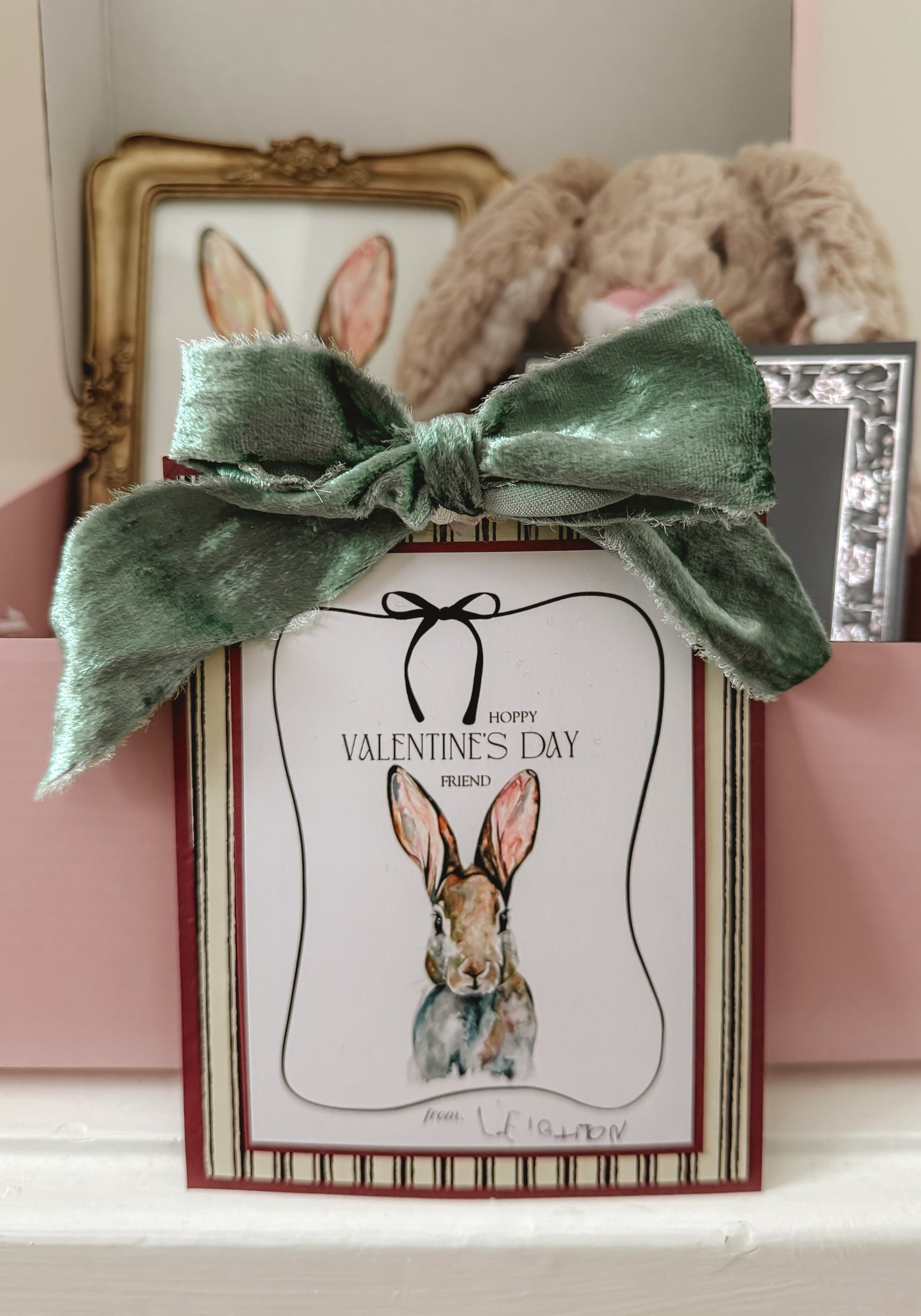 Valentine's Day card with a rabbit illustration and green bow, placed on a surface with a teddy bear and framed picture in the background.