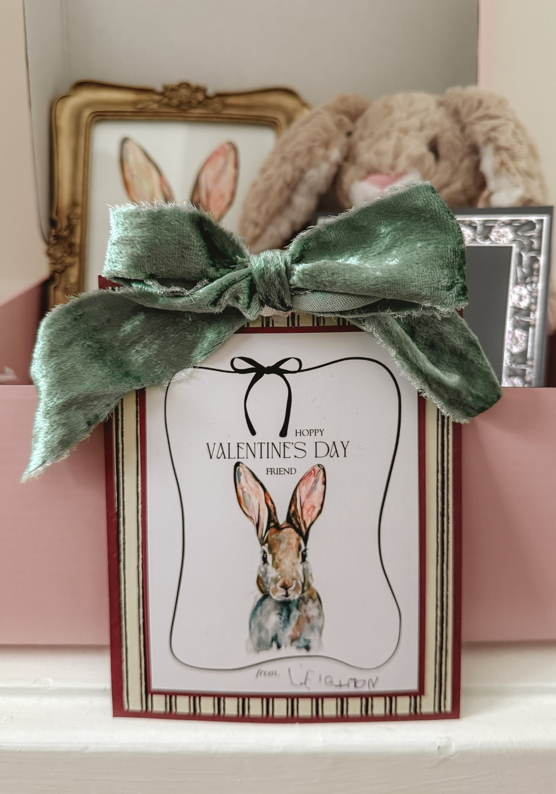 Valentine's Day card with a rabbit illustration and green bow, placed on a surface with a teddy bear and framed picture in the background.